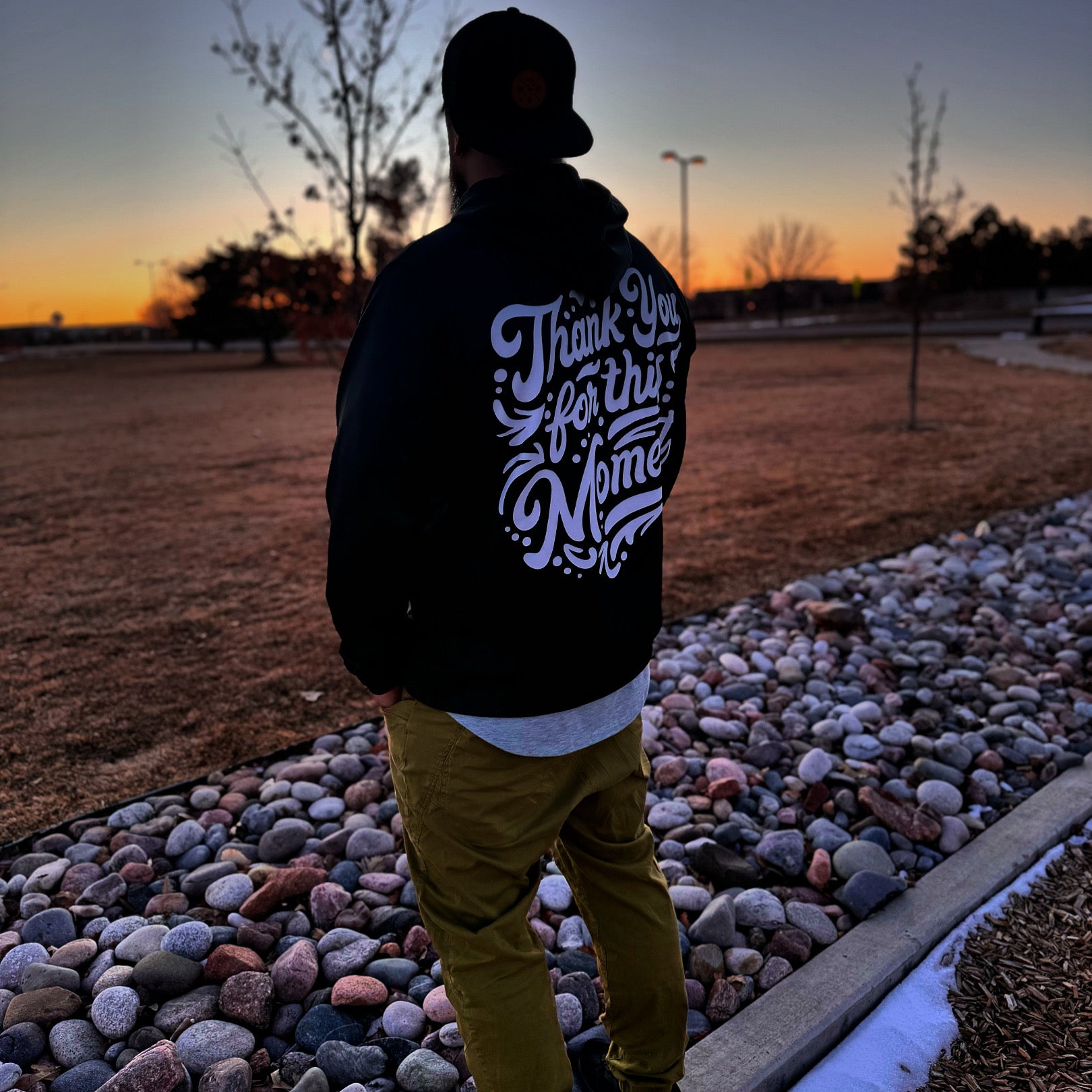 Person wearing a black jacket with white text standing on a pebble path at sunset.